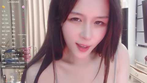 Media: Video of an East Asian woman with long black hair, fair skin, and light makeup, wearing a black top, smiling seductively in a dimly lit room.