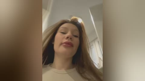 Media: Video of a young woman with long, straight brown hair, fair skin, and closed eyes, wearing a beige top. She stands indoors, with blurred, white-walled background and a mirror reflection.