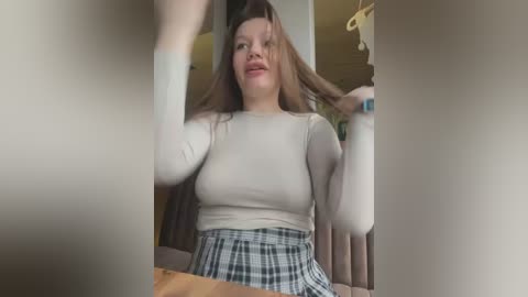 Media: Video of a young Asian woman with long brown hair, wearing a white long-sleeve top and a plaid skirt, adjusting her hair in a living room with a wooden floor and beige furniture.