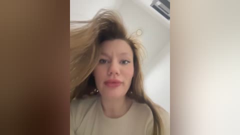 Media: A video of a woman with fair skin and long, blonde hair, captured from a low angle, looking upwards with a neutral expression. The background is blurred, featuring a white bathtub and a window.