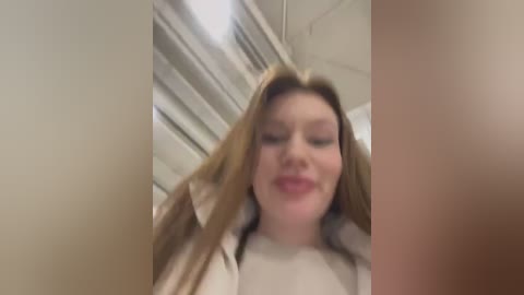 A blurred video of a smiling, light-skinned woman with long, straight blonde hair and light makeup, wearing a white jacket, standing in an indoor setting with beige walls and vertical blinds.