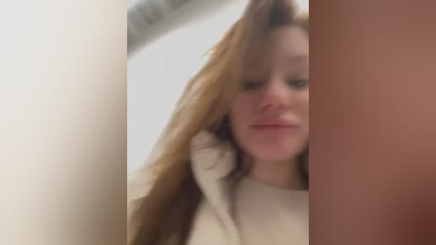 Media: A blurred, close-up video of a young woman with fair skin and long, wavy red hair, wearing a cream-colored sweater. She appears to be touching her hair with her right hand. The background is out of focus.