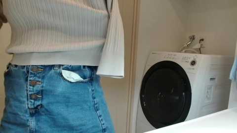 Media: A video showing a person's torso in a white ribbed sweater and blue denim overalls with a white pocket, standing next to a white front-loading washing machine in a laundry room.