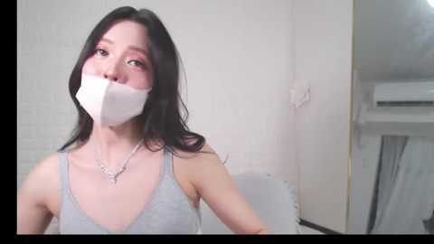 Media: Video of an Asian woman with long black hair, wearing a light gray tank top, gagged with white duct tape, standing in a white room with a window and air conditioner visible.