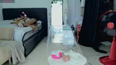 A video of a messy bedroom with a person lying on a bed, surrounded by scattered objects, including a red fan, toys, and a pink vibrator.