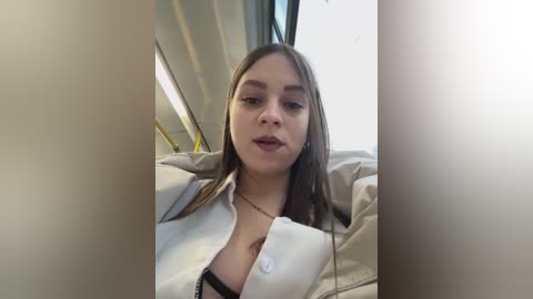 Media: Video of a young woman with straight, light brown hair, fair skin, and medium-sized breasts, wearing a partially unbuttoned white shirt and a black bra, seated in a car with beige upholstery.