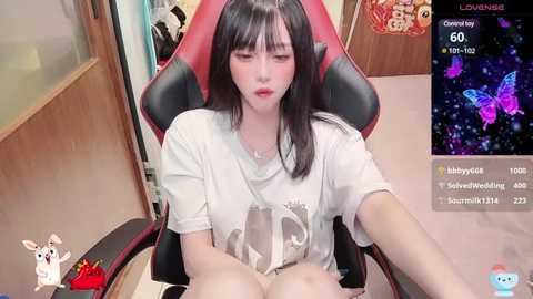 Media: Video of an East Asian woman with long black hair, fair skin, and large breasts, wearing a white T-shirt, seated on a red gaming chair. Background includes a wooden door, a virtual chat screen showing butterflies and text.