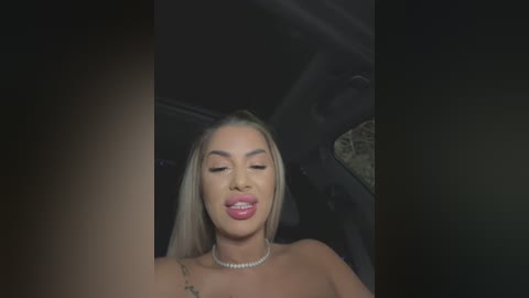 Video of a blonde woman with light skin, wearing a strapless dress, sitting in a car. She has a slim figure, small breasts, and is smiling with her eyes closed.