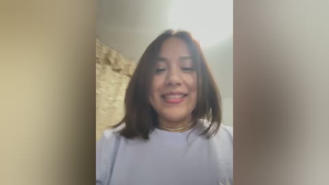 Media: Video of a smiling Latina woman with medium skin tone, shoulder-length dark brown hair, wearing a white top, standing against a beige wall with a stone texture.