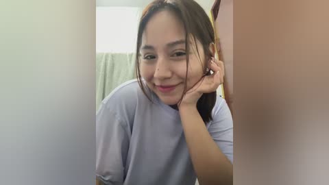 Media: Video of a young Asian woman with straight black hair, wearing a light blue shirt, smiling, leaning on a sink in a bathroom with beige walls and a towel hanging on the left.