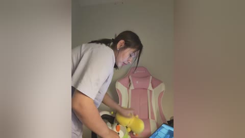 Media: A video of a young woman with brown hair tied back, wearing a light grey T-shirt, playing a video game in a dimly lit room.