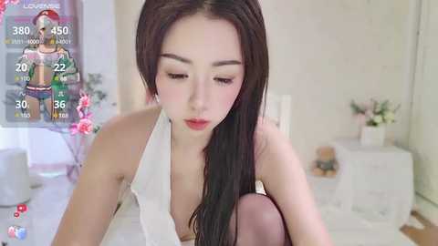 Media: A video of a young East Asian woman with long black hair, wearing a white halter top, sitting in a bedroom. Background includes a white dresser, bed, and a poster of a cartoon character.