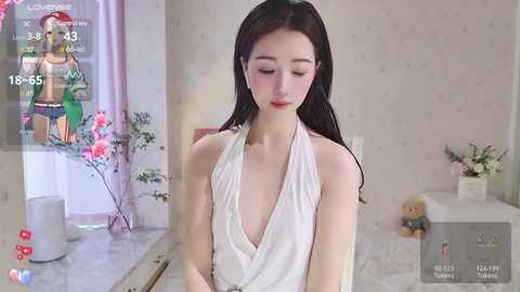 Media: A video of an Asian woman with long black hair, wearing a white halterneck dress, standing in a softly lit bedroom. The room features light-colored walls, a bed, and a pink flower arrangement.