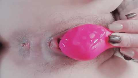 A close-up video shows a fair-skinned person's vulva being penetrated with a bright pink, shiny silicone anal plug. The person's hand with metallic nail polish holds the plug.