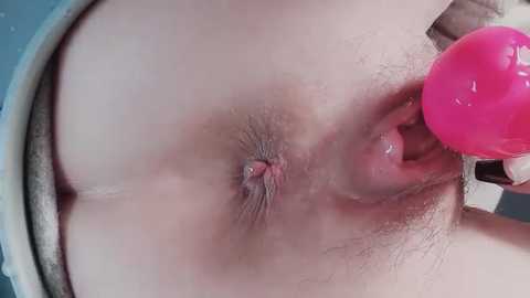 Close-up video of a nude person's anus, showing a pink anal plug inserted and held by a hand. Background shows a white, textured surface.