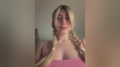 Media: Video of a young, fair-skinned woman with long blonde braids, wearing a pink tank top, standing in a minimalist room with beige walls.