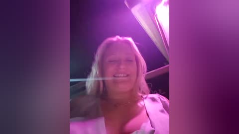 Media: Video of a smiling, fair-skinned blonde woman with medium-length hair, wearing a white shirt, sitting in a dimly lit car with pink and purple neon lights.