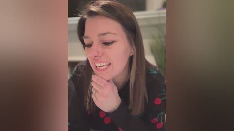 Media: Video of a young woman with straight, brown hair, wearing a black floral-patterned sweater, smiling and holding a phone. The background is blurred, showing a white window sill.