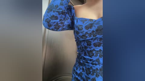 Media: Video of a woman in a blue floral dress, taken from a side angle, showing her torso and arm. She is in a room with a metallic background and a blue wall.