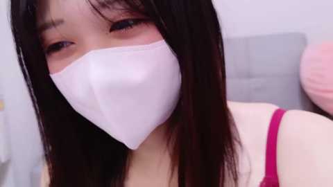 Video of an Asian woman with long black hair, wearing a white face mask and a pink spaghetti-strap top, looking slightly to the side. Background includes a grey couch and a pink pillow.