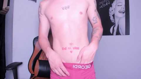 Media: Video of a shirtless, slender, fair-skinned man with tattoos, wearing red Calvin Klein underwear, standing in a room with Marilyn Monroe posters and a black chair.