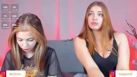 Media: Video of two young women with fair skin and long, straight brown hair. One wears a black sheer top, the other a black tank top. They sit on a couch in a dimly lit room with pink lighting.