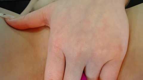 Media: Close-up video of a light-skinned hand with slightly red skin, fingers spread, touching a pink object, possibly a vibrator, against a blurred dark background.