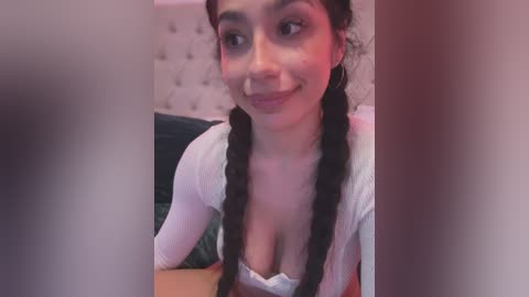 Media: A video of a young woman with light skin, long black hair in braids, wearing a white top with a low-cut neckline, smiling. She's indoors, on a bed with a beige headboard.