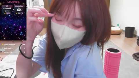 Media: A video of a woman with long brown hair, wearing a face mask, adjusting her glasses in a room with a wooden table, pink mug, and a black cup.