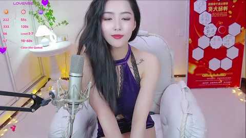 Media: Video of an Asian woman with long black hair, wearing a purple halter top and white pants, sitting on a white chair. She has a nose ring and is singing into a microphone. Background includes a red poster and a plant.