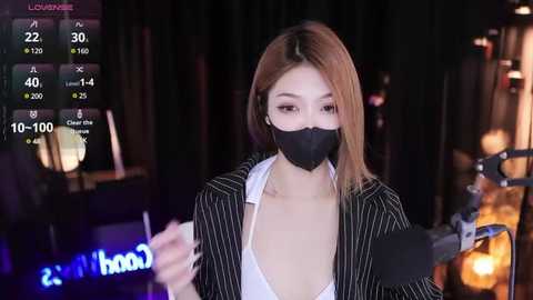 Media: A video captures an East Asian woman with straight, light brown hair, wearing a black face mask, pinstripe blazer, and white blouse, speaking into a microphone.