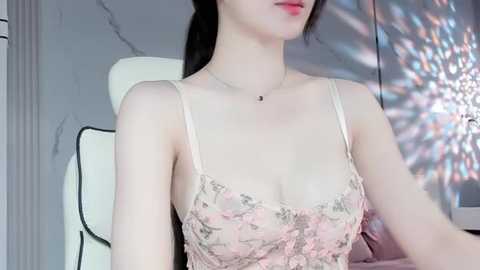 Media: Video of a fair-skinned woman with long black hair, wearing a sheer pink lace camisole with floral embroidery. She is seated indoors with a white chair and a blurred background featuring a light-colored wall and a window with colorful light patterns.