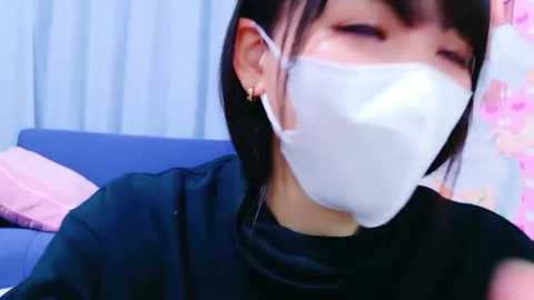 Media: A video of an Asian woman with short black hair, wearing a white face mask, a black shirt, and small earrings, indoors against a blue couch and pink floral wallpaper.