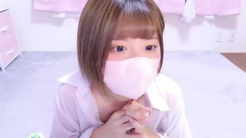 Media: Video of a young Asian woman with shoulder-length brown hair, wearing a white medical mask, kneeling on a white tiled floor. She's holding a pair of hands. The background shows a pink curtain and a white dresser.