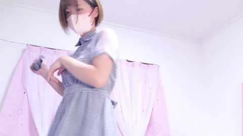 Media: A video of a young woman with short, light brown hair, wearing a sheer, light blue dress, standing in front of a white wall with pink curtains. She has a mask on and appears to be moving slightly.