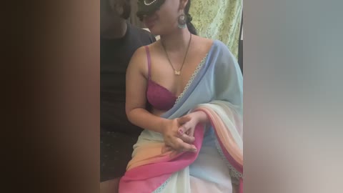 Media: Video of a South Asian woman with medium skin tone, wearing a purple bra and a colorful sari with a white and pastel gradient, sitting on a dark couch.