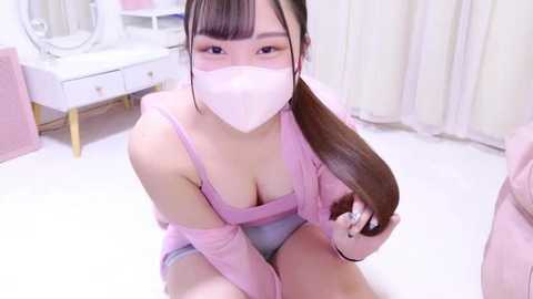 Media: Video of an Asian woman with light skin, long straight brown hair, wearing a pink mask, pink top, and grey panties, sitting on a white floor, holding her hair, in a softly lit room with white furniture.