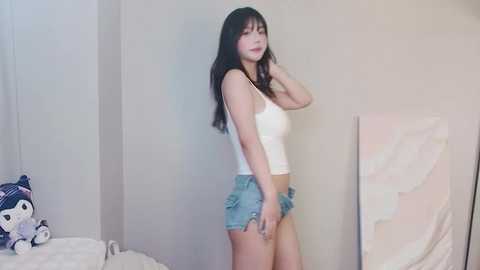 Media: Video of an Asian woman with long black hair in a white tank top and denim shorts, standing in a minimalist bedroom with pastel decor.