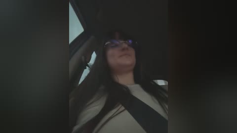 Media: Video of a person with long black hair, wearing glasses and a white shirt, seated in a car with a black seatbelt across their chest, blurry background.