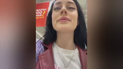 Media: Video of a young woman with long black hair, fair skin, and closed eyes, smiling. She wears a red leather jacket over a white shirt, in a grocery store background.