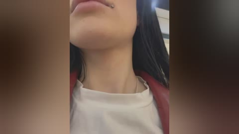 A close-up video of a woman's face and neck, showing her light skin, long black hair, and a small nose piercing. She wears a white t-shirt under a red jacket. The background is blurred, suggesting an indoor setting.