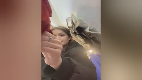 Media: A video of a young woman with long, straight brown hair, wearing a black coat, taking a selfie in a dimly lit room with a reflective surface. She holds a smartphone with a case in her left hand.