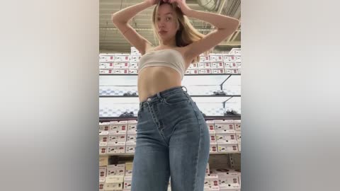 Media: Video of a slim, light-skinned young woman with long blonde hair, wearing a beige sports bra and high-waisted blue jeans, standing in a warehouse with stacks of cardboard boxes in the background.