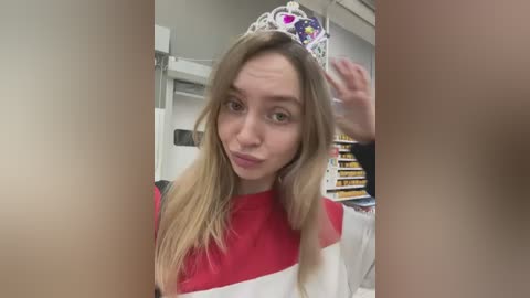 Media: Video of a young woman with long blonde hair, wearing a red and white striped top and a crown, playfully blowing a kiss to the camera in a brightly lit room with shelves in the background.