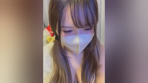 Media: Video of an Asian woman with long, straight brown hair, wearing a white surgical mask, looking down, in a room with a fluffy white rug and a red and yellow plush toy.