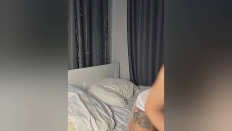 Media: A video of a dimly lit bedroom with a bed, white pillows, gray curtains, and a partially visible tattooed arm. The room is simple and modern.