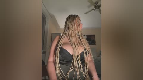 Video of a woman with long, braided blonde hair, wearing a black bra, standing in a dimly lit bedroom with beige walls and a ceiling fan.