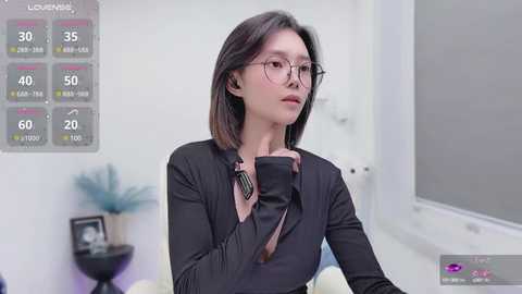 Media: Video of an Asian woman with straight dark hair, wearing glasses, adjusting a black shirt in a modern, minimalist room with a weather display.