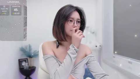 Media: Video of an Asian woman with straight, shoulder-length black hair, wearing glasses, a white off-shoulder top, and pensive expression. Background features a modern, minimalistic room with a window and decorative items.