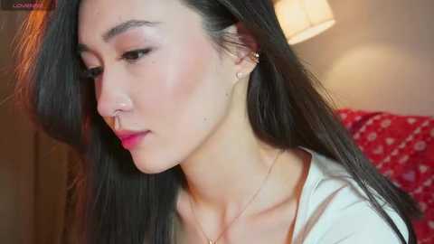 Video of an Asian woman with long black hair, wearing a nose ring and a white top, in a softly lit room with a red-patterned cushion and a lamp.